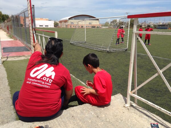 R58691 N No Restrictions Om Worker Tying The Shoelaces Of A Young Refugee Boy Before His Football Game.